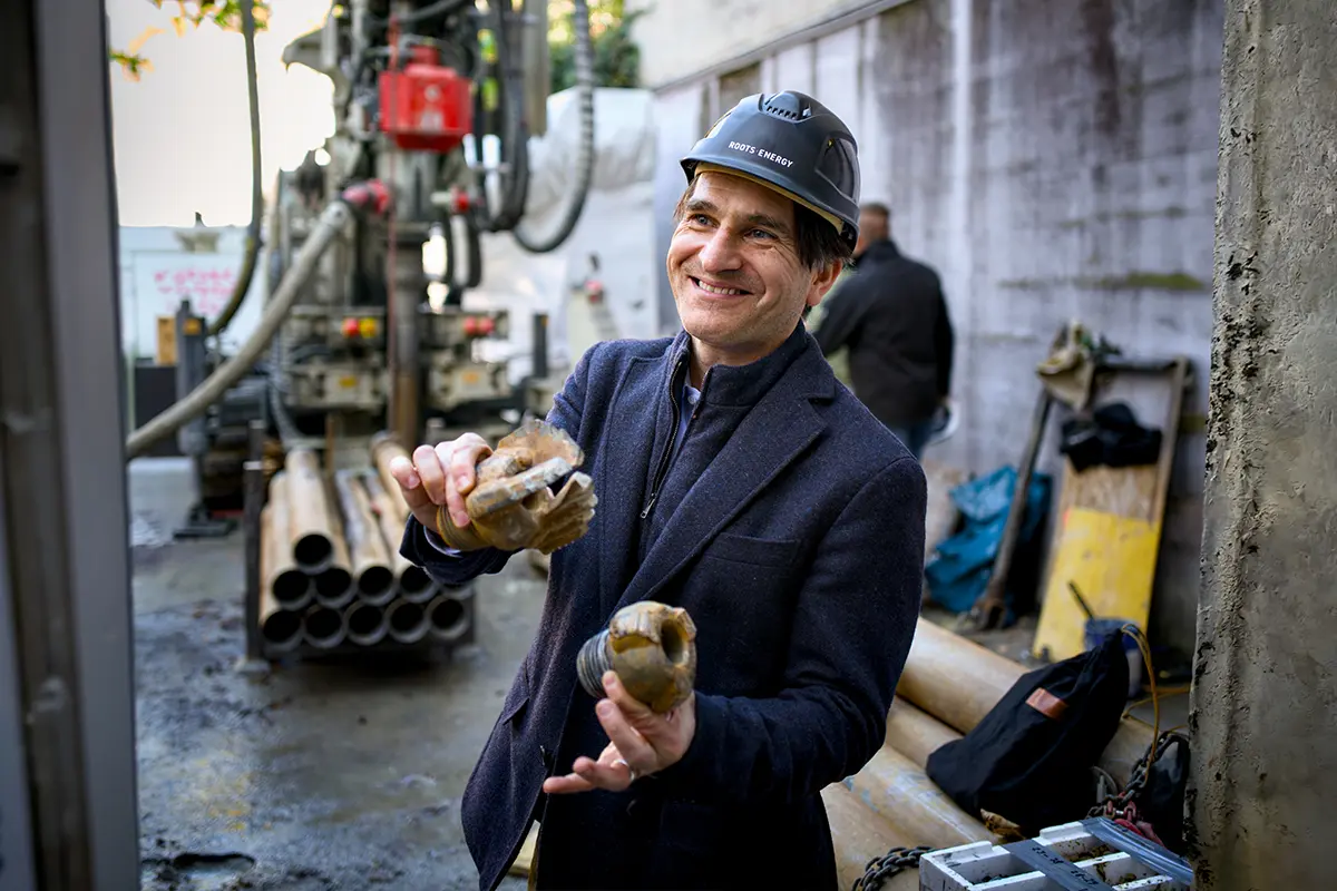 Person in a Roots·Energy helmet holding drill bits in front of the drilling rig